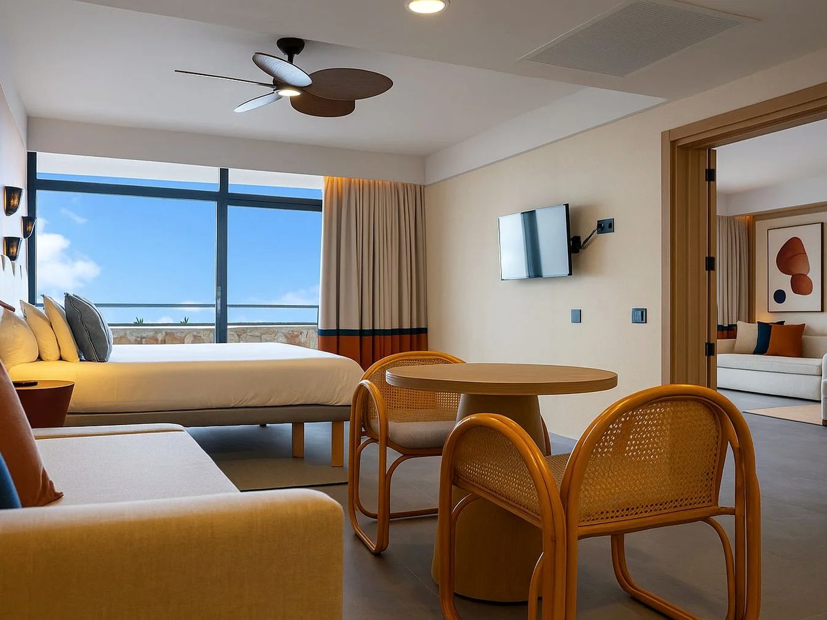Open-plan hotel suite with a double bed, a sitting area with a round table and rattan chairs, and a doorway through to a second room.