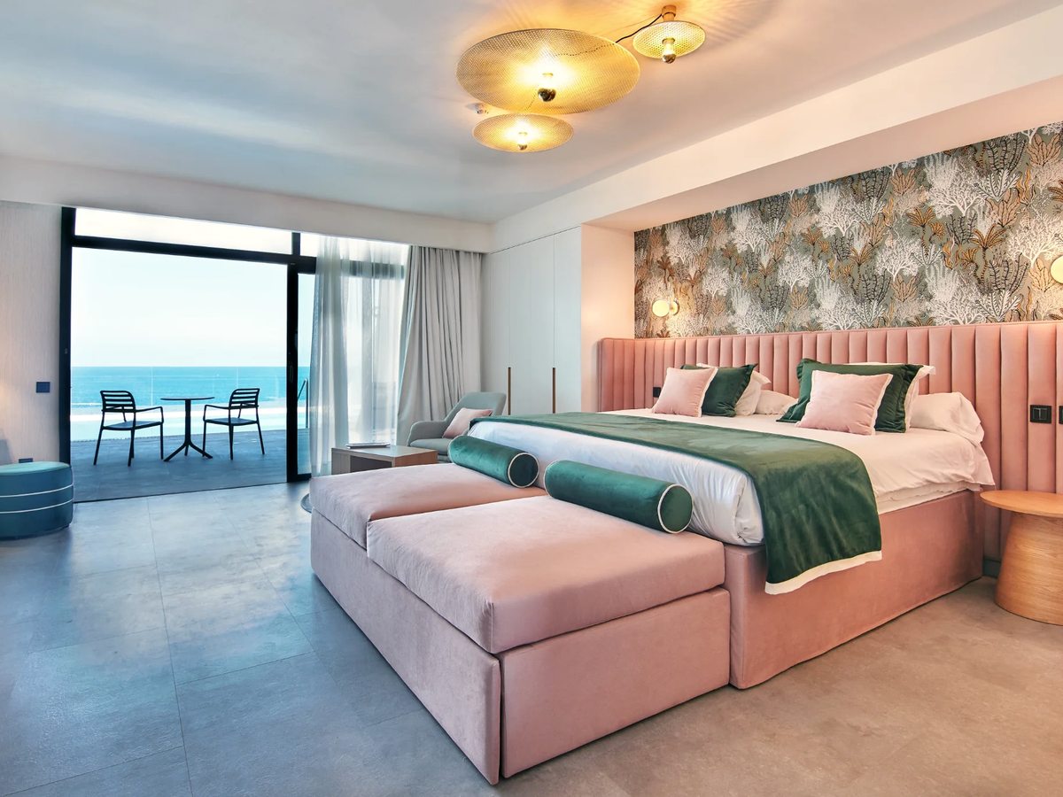 Hotel room with a pink upholstered bed and a botanical-print wall, sliding doors open onto a furnished terrace above the sea.