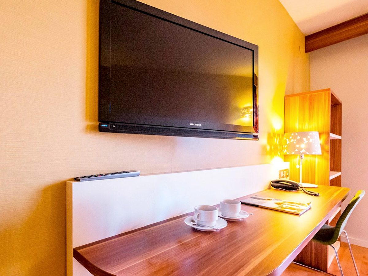 A wall-mounted flat-screen TV above a long wooden desk holding two coffee cups on saucers, a table lamp and a telephone in a suite