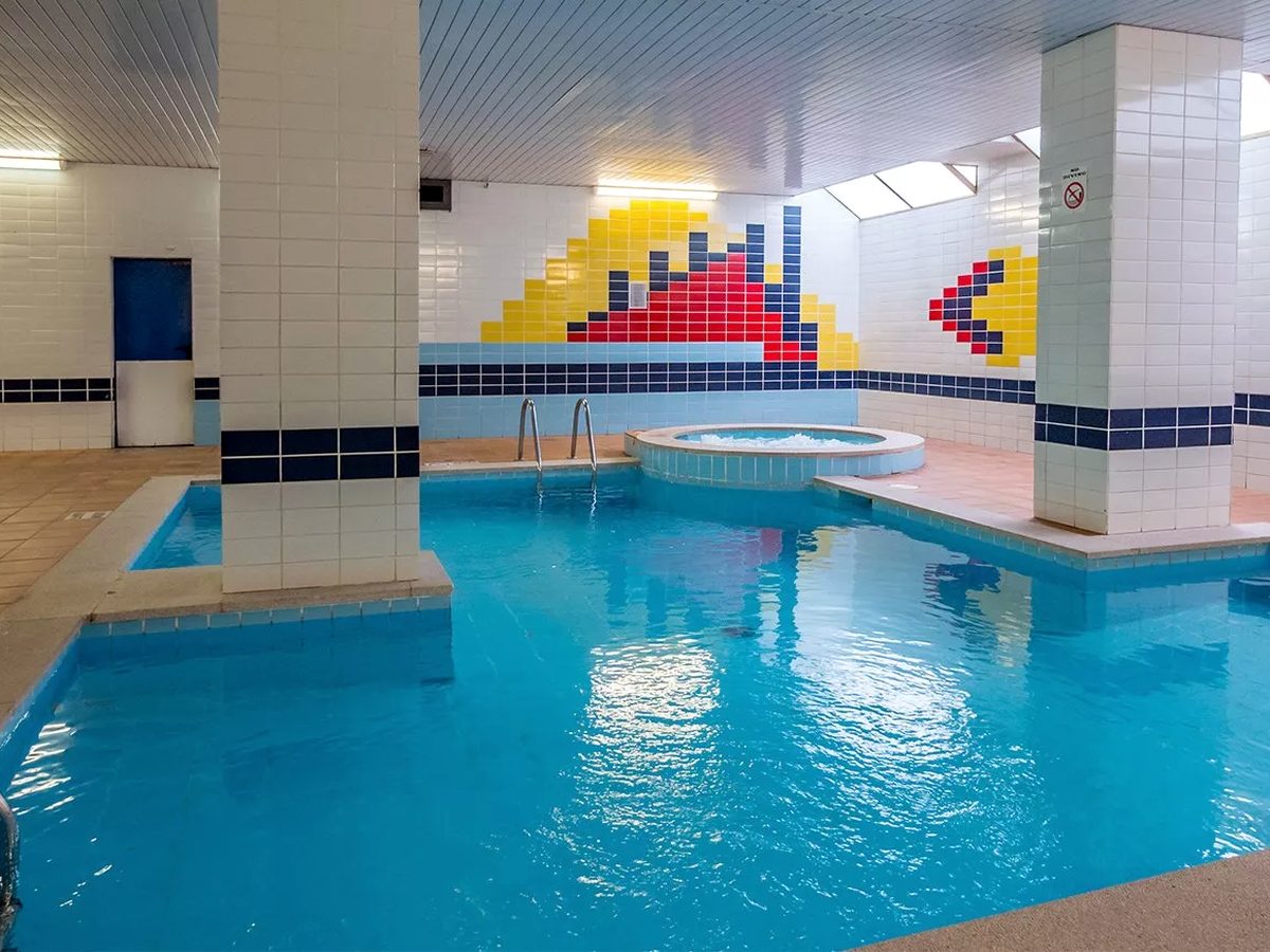 An indoor pool with clear blue water, a round jacuzzi set into one side, white-tiled columns and a bright geometric tile mural along the wall.