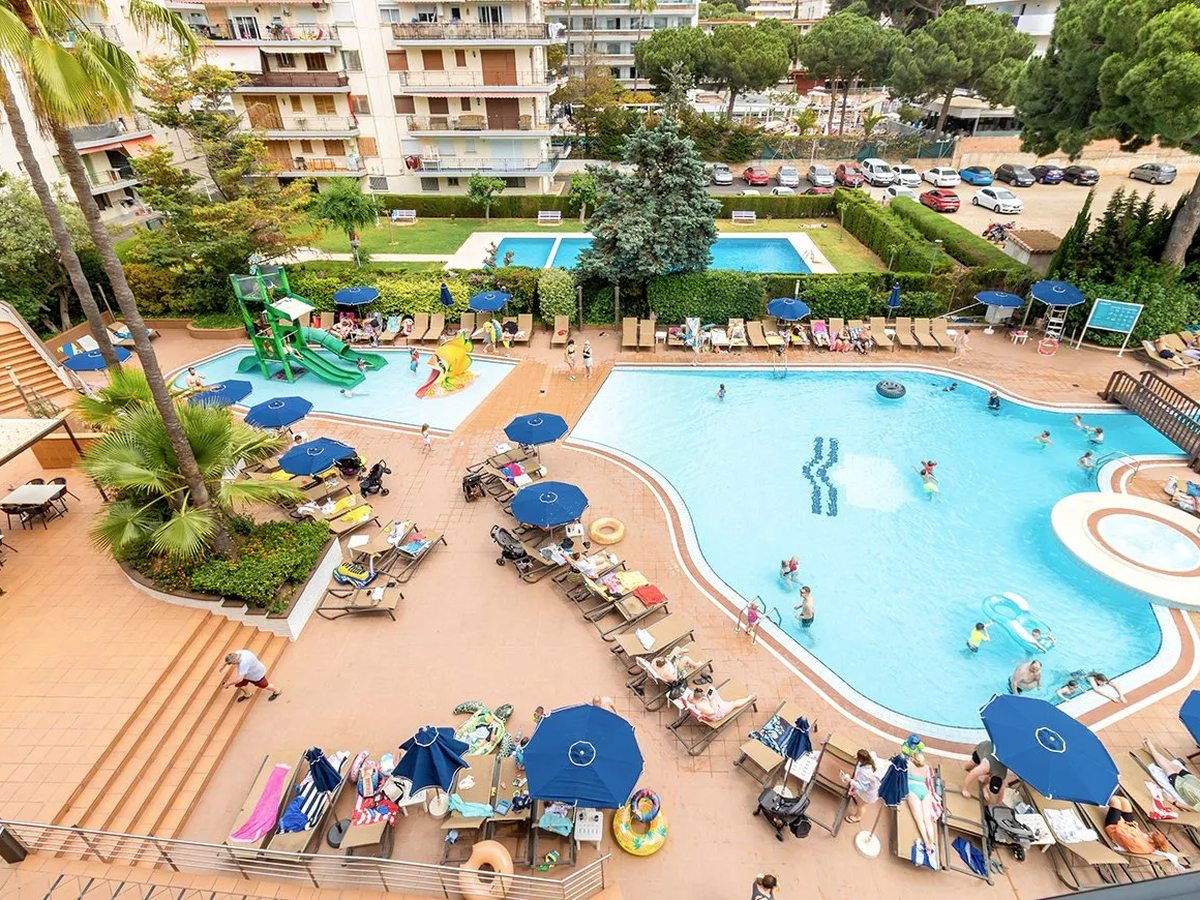 An aerial view of the pool complex: a children's pool with green slides on the left, the freeform main pool with a round jacuzzi on the right, and a rectangular pool behind a hedge, all edged with loungers and blue umbrellas.