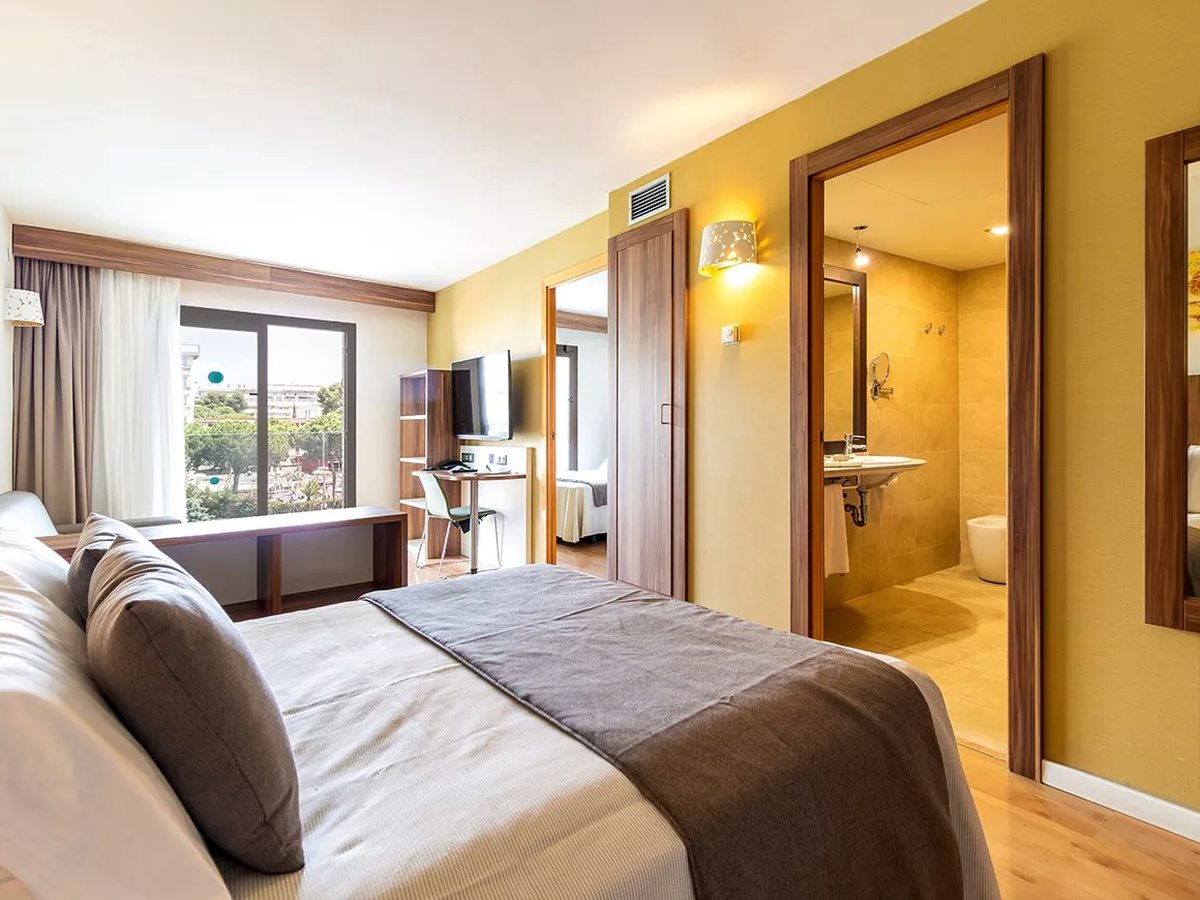 A double bedroom with a grey throw and a wall-mounted TV, a window over the town, and open doors to a second bedroom and an ensuite bathroom.