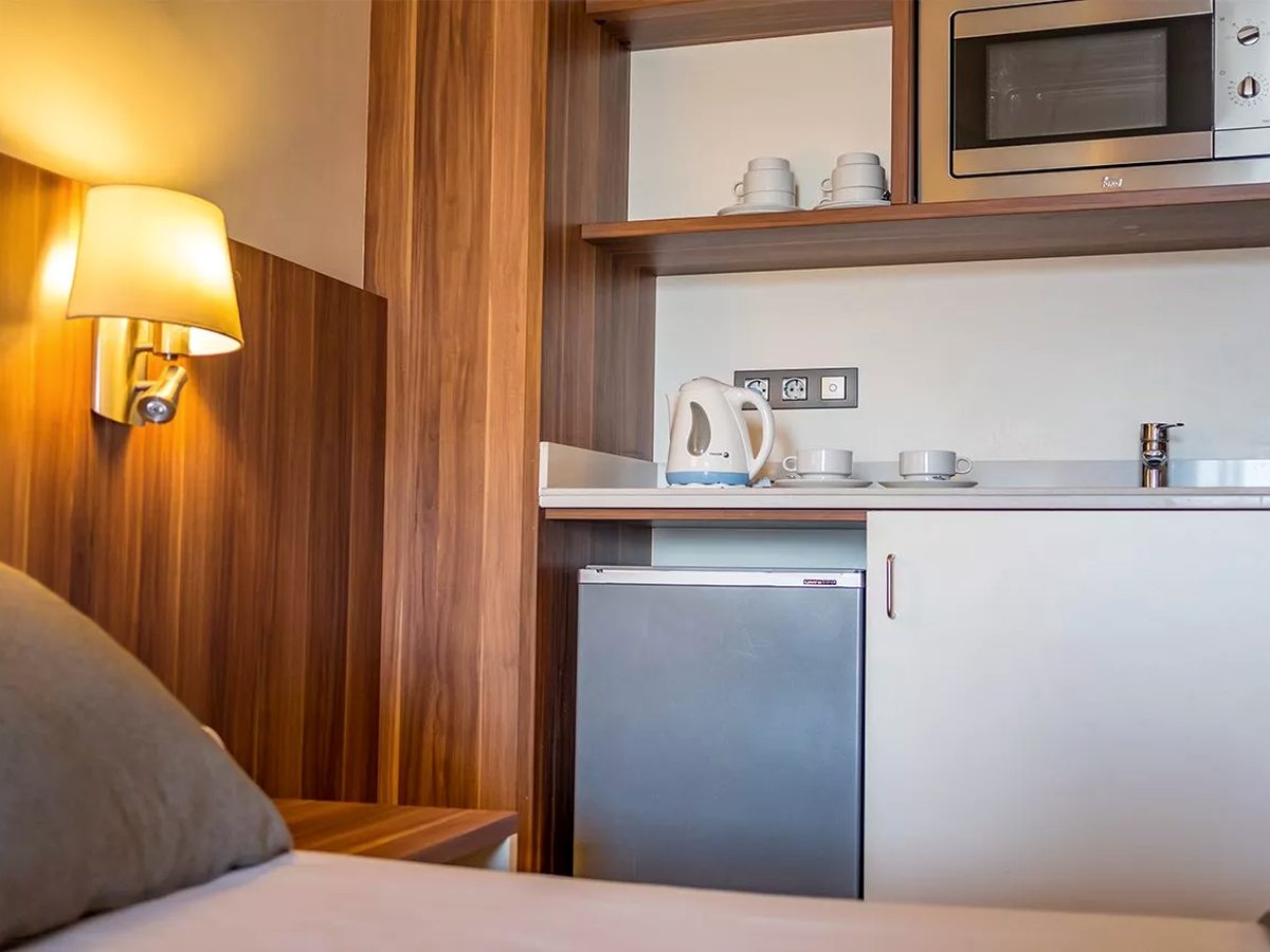 A compact in-room kitchenette with a microwave on a shelf, an electric kettle and cups, a sink and an under-counter fridge, beside the bed.
