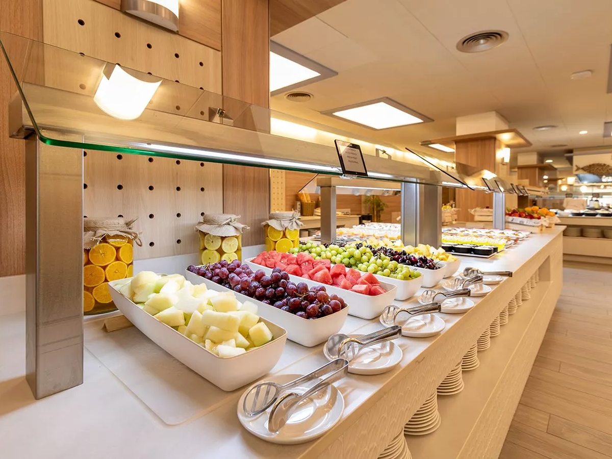 A long buffet island lined with bowls of fresh fruit, salads and cold dishes on white platters, with stacked plates at the end.