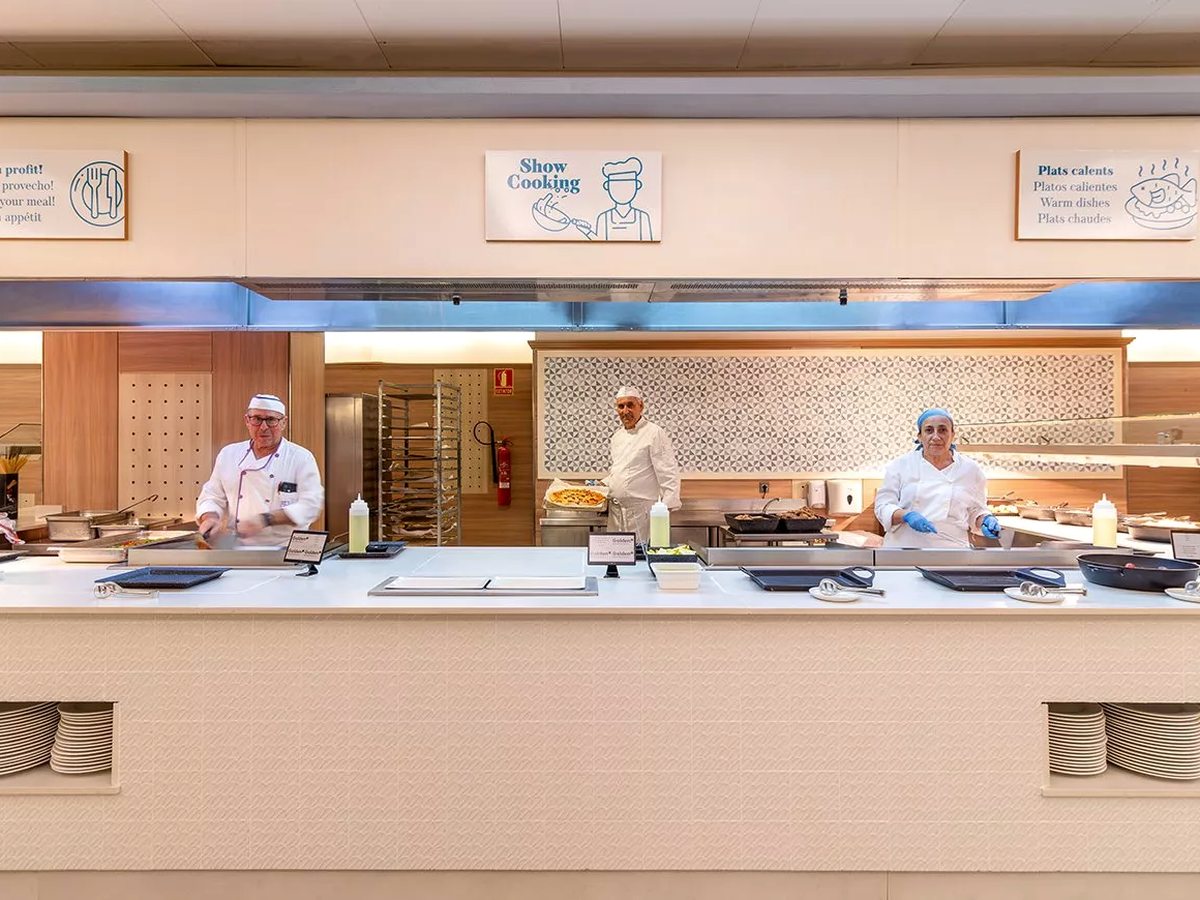 Three chefs in whites working at a live show-cooking counter behind a low glass screen, with a menu board above.