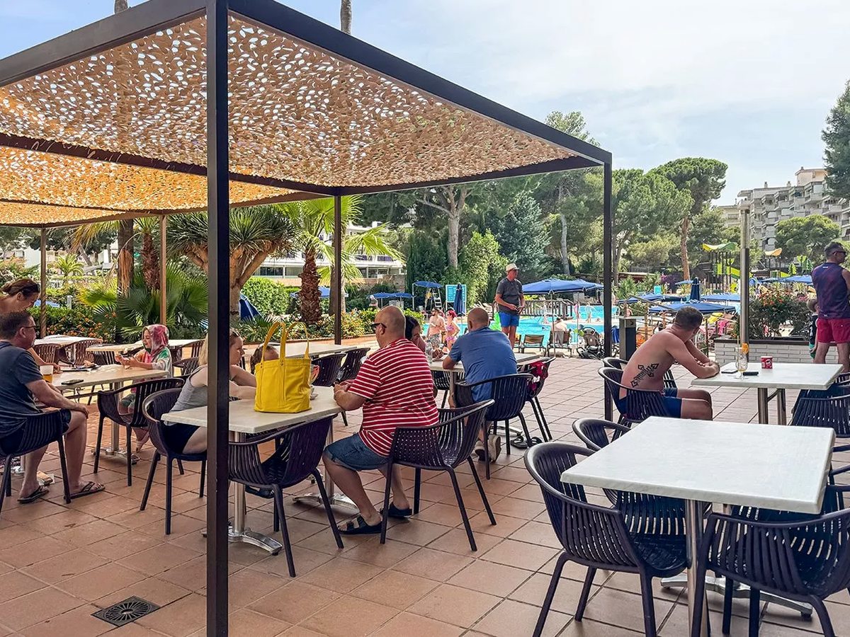 An outdoor dining terrace under a patterned pergola canopy with dark chairs at white tables, families seated, and the pool and gardens behind.