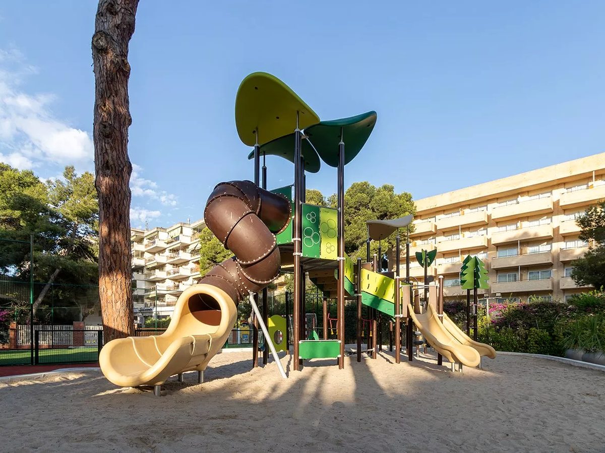 An outdoor playground with a tall climbing-and-slide structure under a canopy, set on sand behind a wall, with pine trees and the hotel block behind.