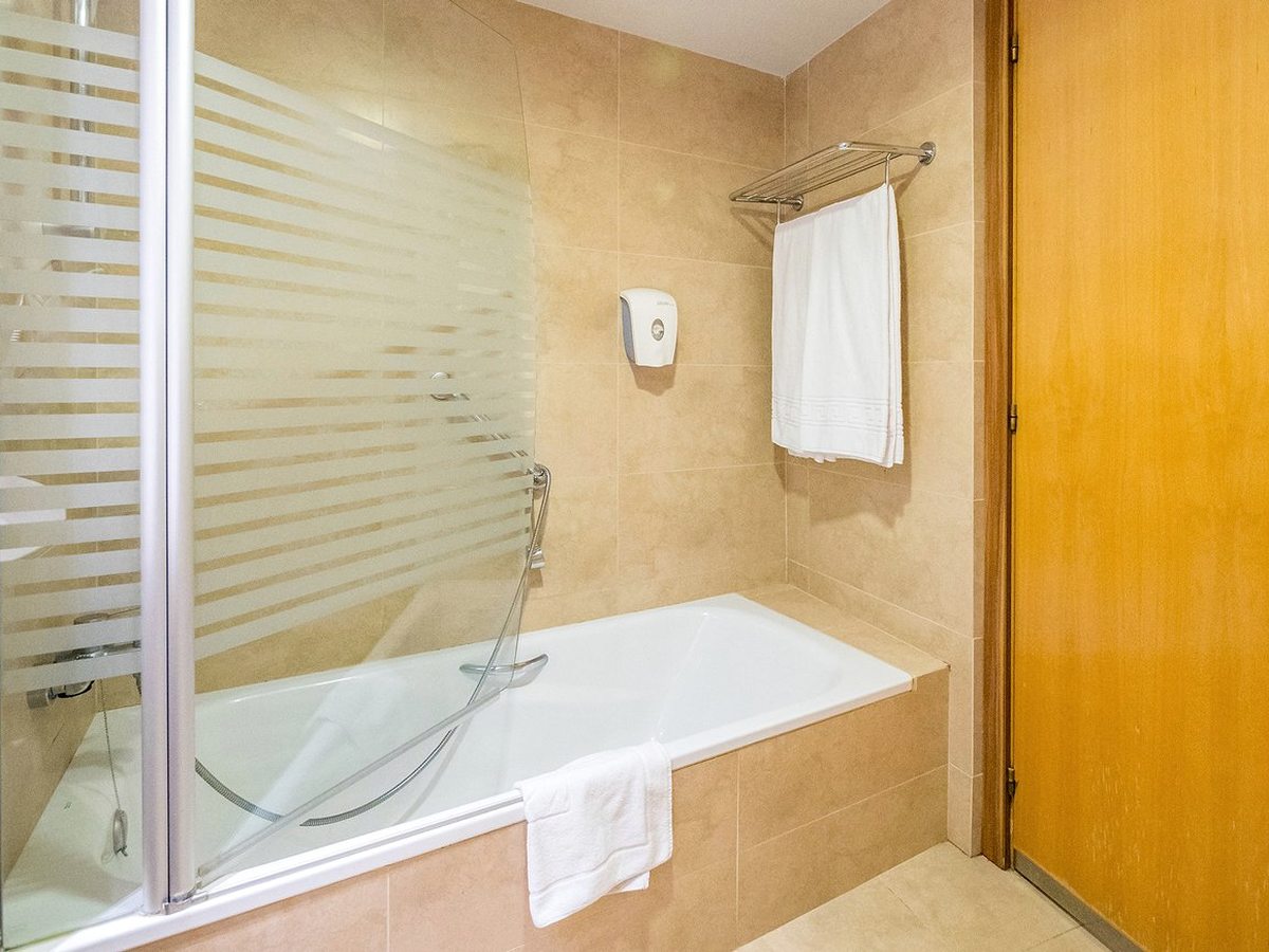A tiled bathroom with a bath, a folding glass shower screen over it, a hand shower and towels on a rail.
