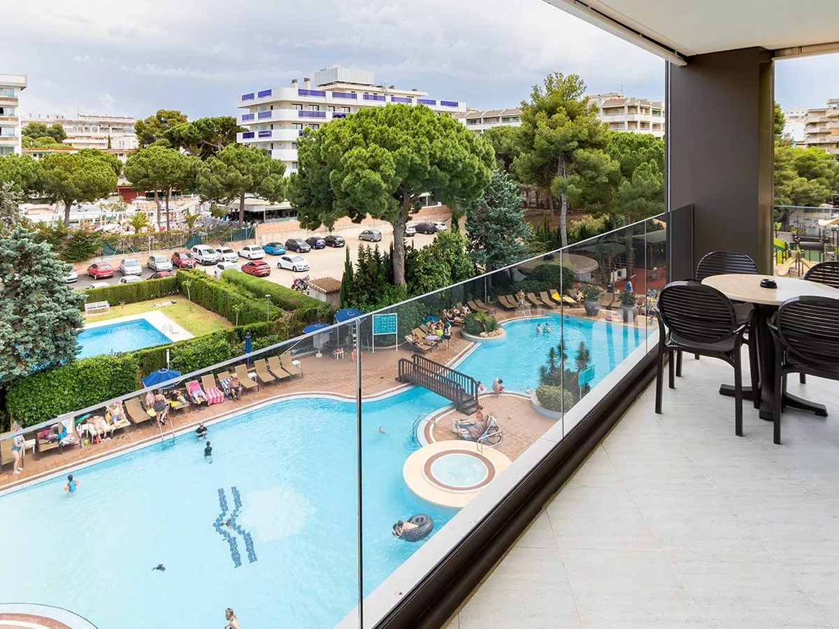 View from a suite balcony over the main pool with a round jacuzzi and a smaller rectangular pool, edged with sun loungers and gardens, with apartment blocks and parked cars beyond.