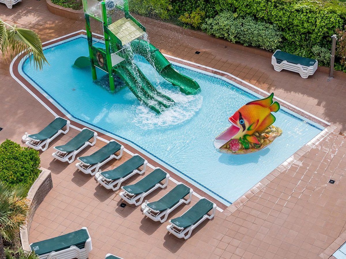 A shallow children's pool with green tube slides and a tipping structure, a painted open-mouthed fish figure at one end, and green sun loungers along the tiled surround.