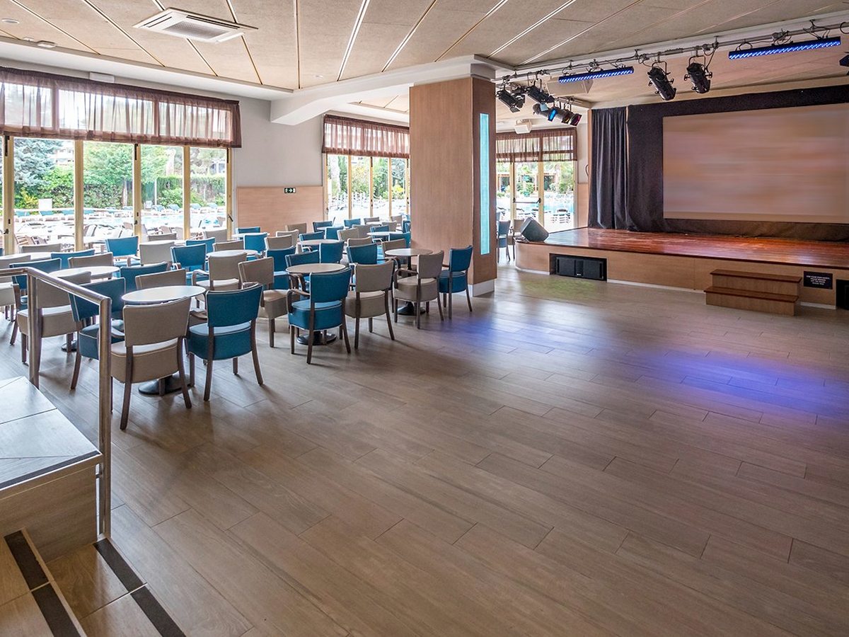An indoor entertainment room with a raised wooden stage, a large screen, stage lighting rigs, and blue and cream tub chairs at small round tables by garden windows.
