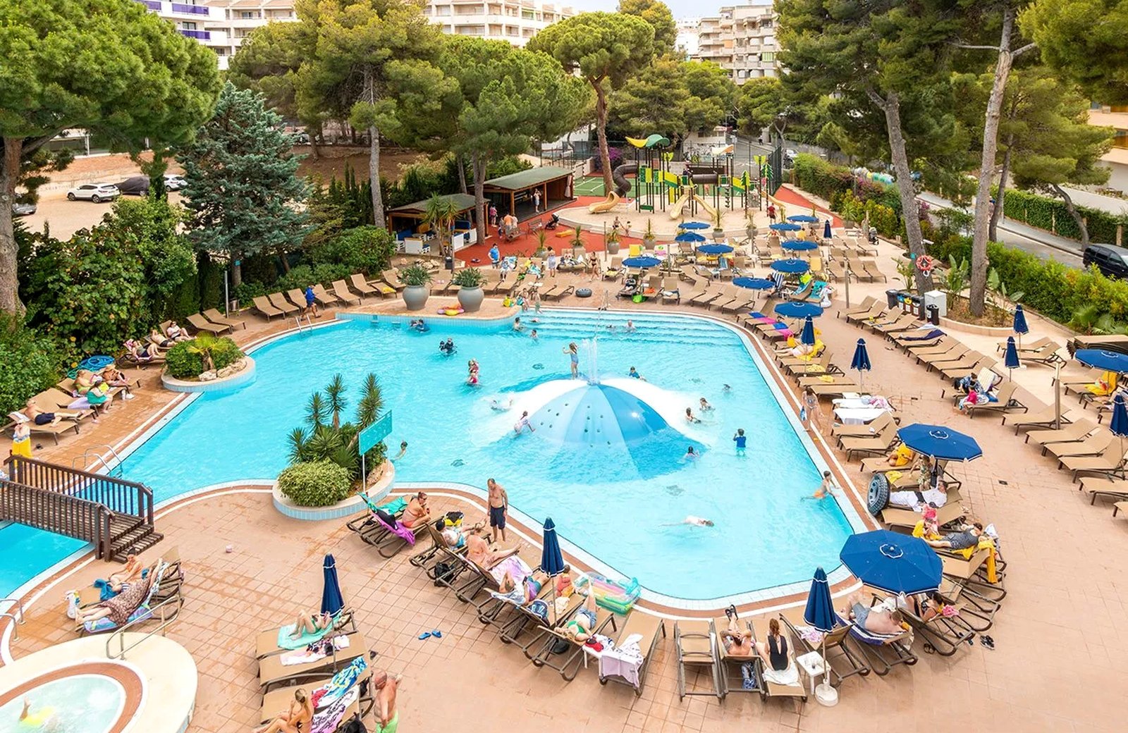 The main outdoor pool at Golden Avenida Family Suites, ringed with sun loungers and umbrellas, a domed water feature in the middle and the playground behind.