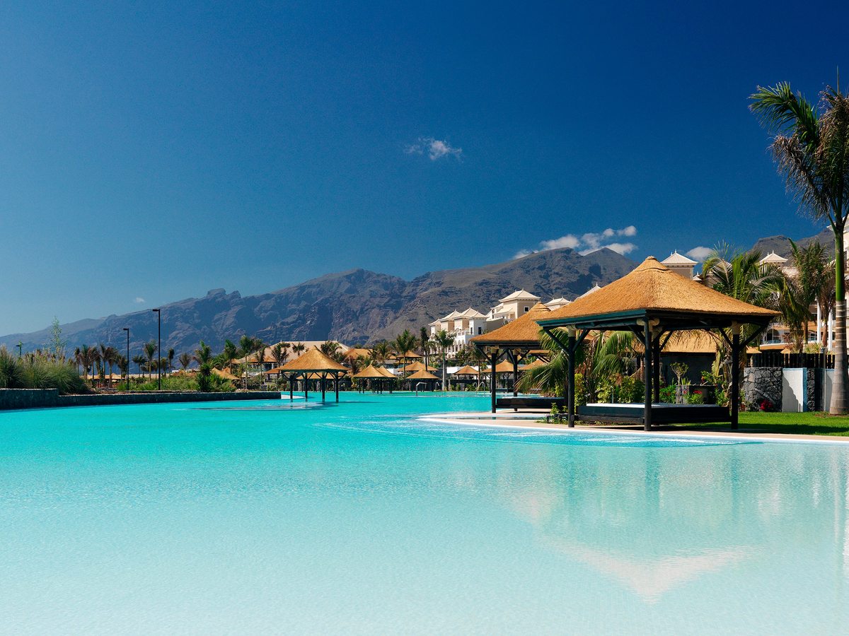A large pool with thatched pavilions set in the water and palms around it, a range of mountains behind under a deep blue sky.