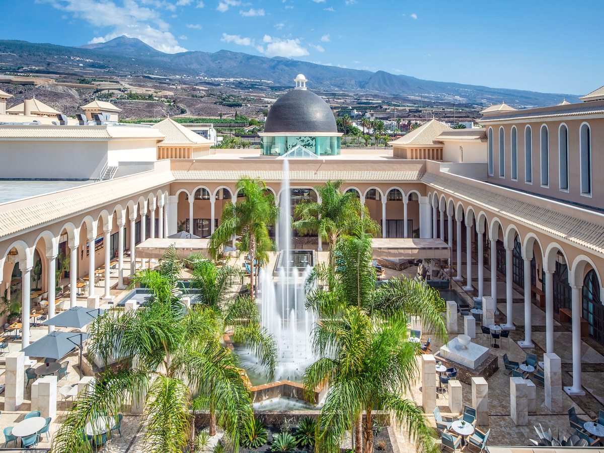 A large arcaded courtyard with white colonnades around a tall central fountain, palm trees and a domed roof beyond, mountains in the distance.