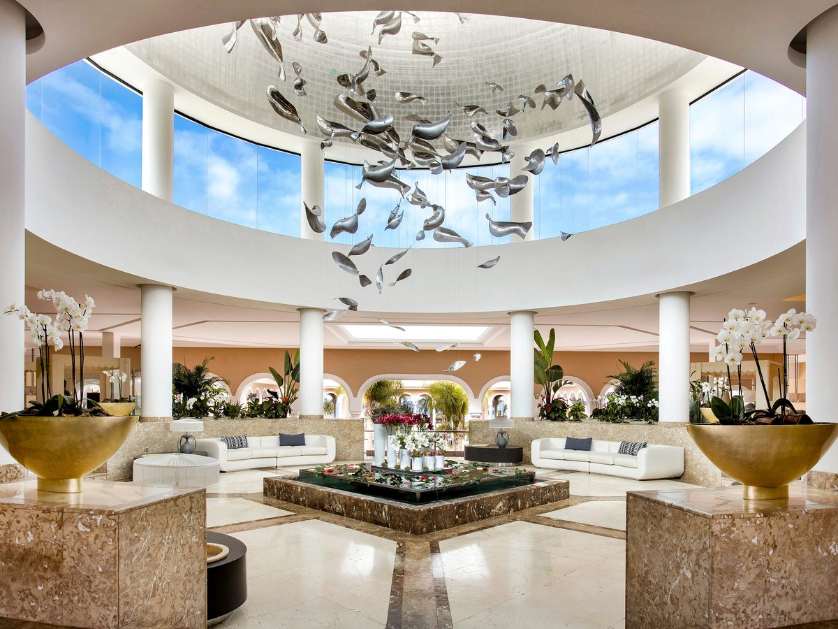 A circular lobby beneath a domed skylight, with a suspended metal shoal sculpture, white columns, a marble floor and white sofas.
