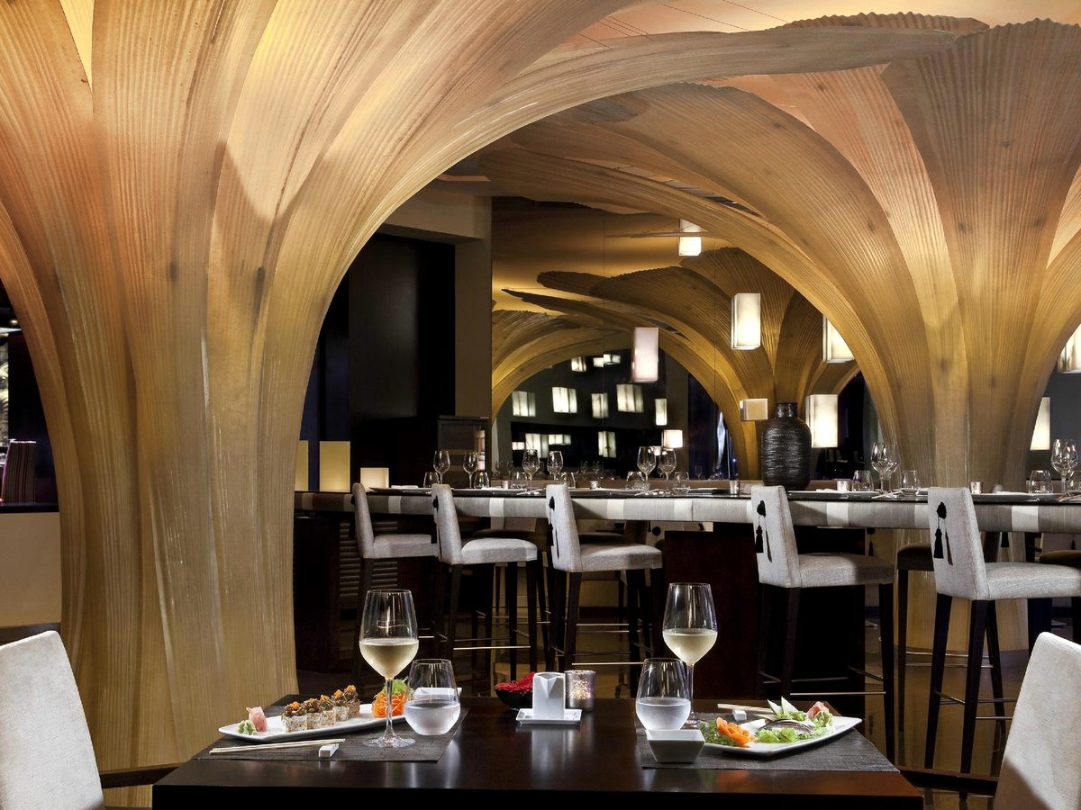 A restaurant with a sculptural pale-timber vaulted ceiling, a bar counter with stools and tables set with sushi and white wine.
