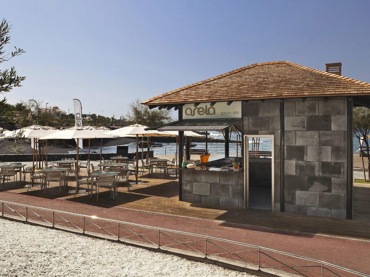 A single-storey stone beach bar with a shaded terrace and white parasols on a paved seafront promenade.