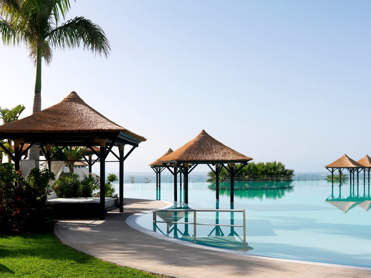 A calm infinity pool edged with lawn and thatched gazebos, the sea on the horizon.