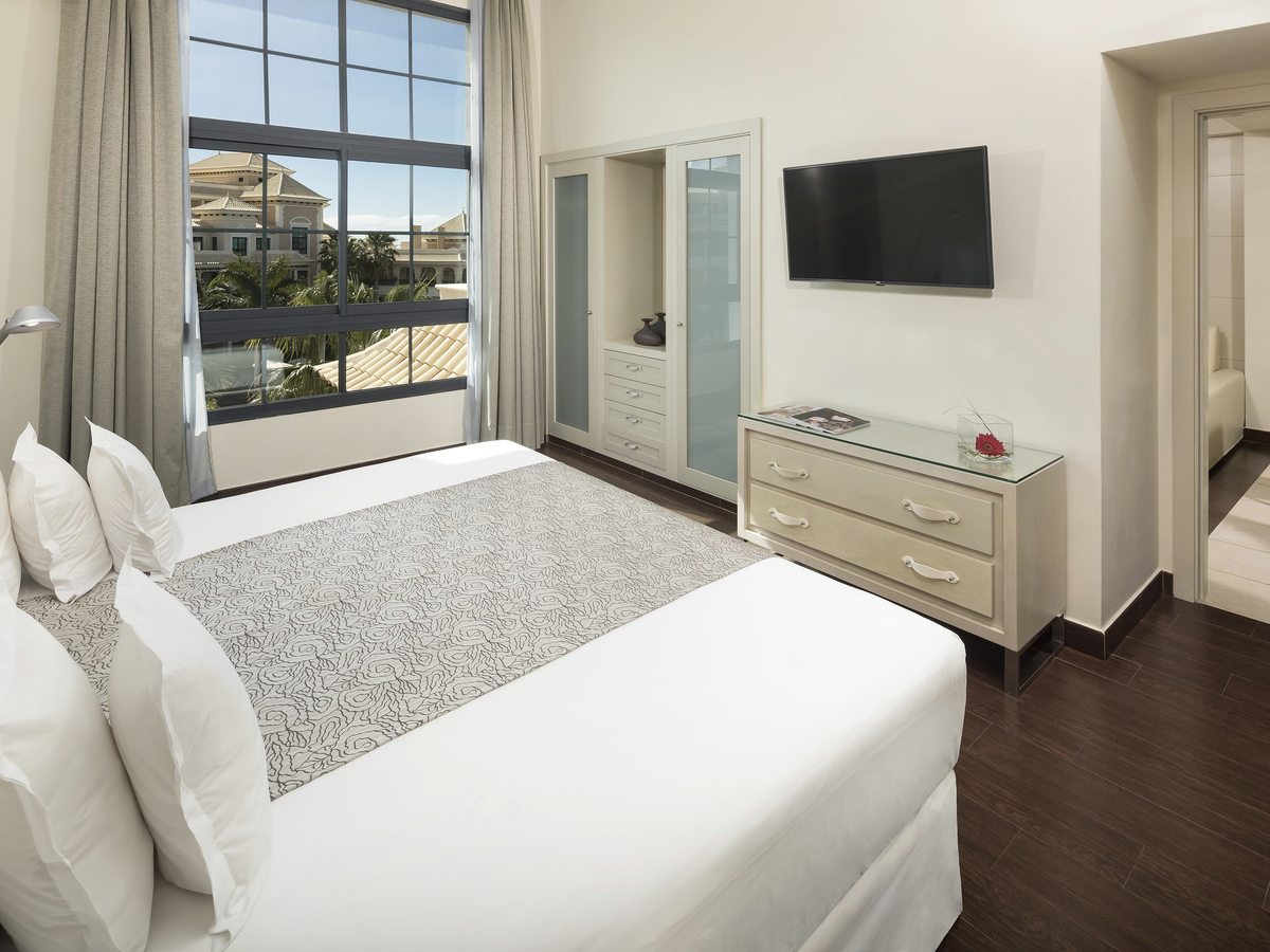 A bedroom with a double bed and white bedding, a wall-mounted TV, a chest of drawers and a tall window.