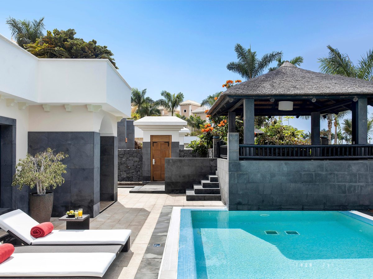 A private plunge pool on a stone terrace with twin loungers, red towels, a shaded gazebo and white villa walls.
