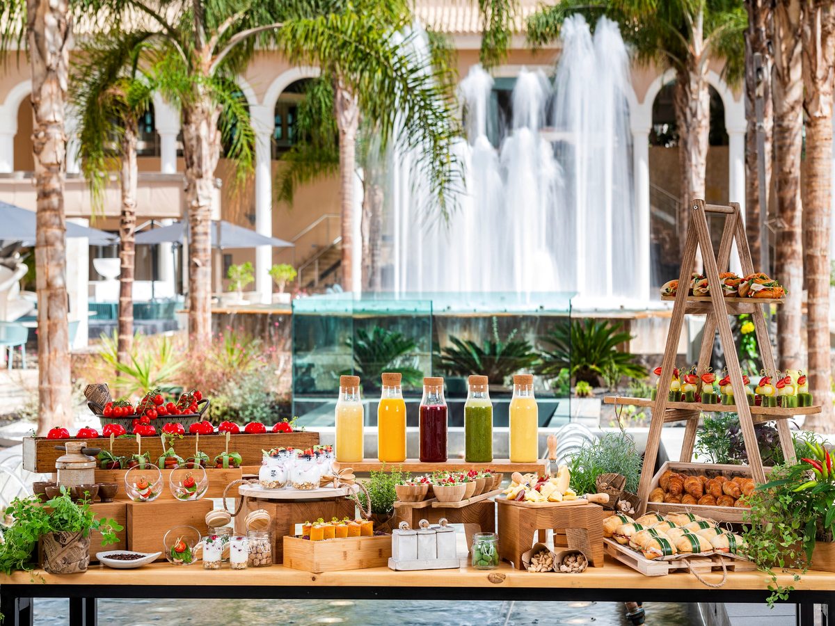 A wooden table laid with bottled juices, tomatoes, pastries, granola jars and small dishes, a courtyard fountain playing behind it.