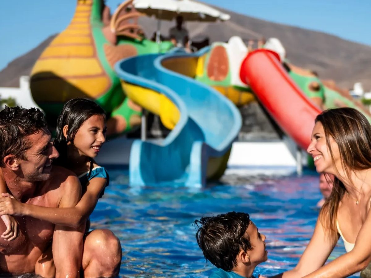 Children and parents in a shallow pool in front of a large green dragon water-play structure with curling blue and red slides.