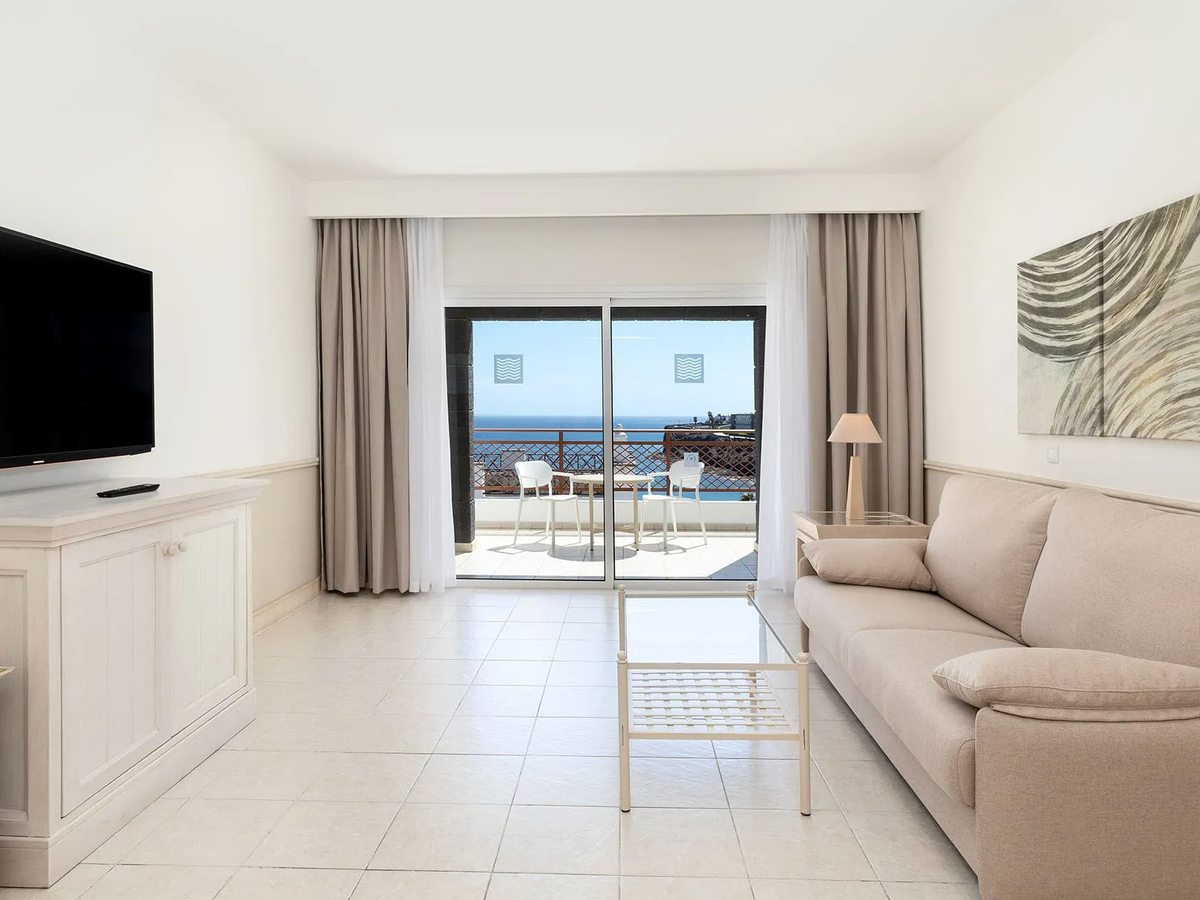 Suite living area with a beige sofa, wall-mounted TV and coffee table on a white tiled floor, with sliding doors to a sea-view terrace.