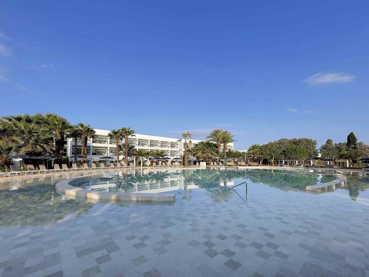 Large freeform pool with a wide tiled shallow entry, sun loungers and palms reflected in the water, the resort buildings behind.