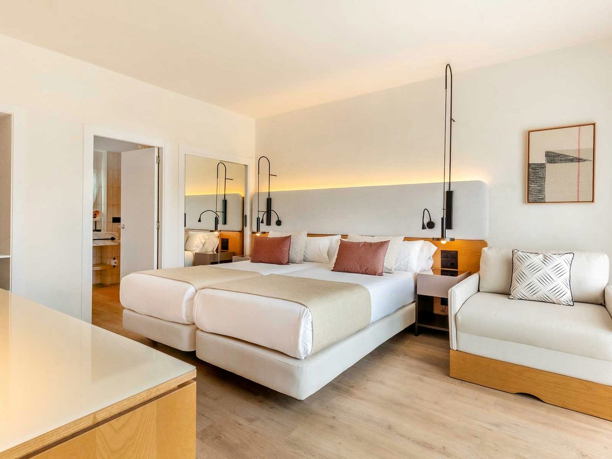 Room with two single beds pushed together, bedside tables, pendant reading lights and a single sofa against a cream wall.