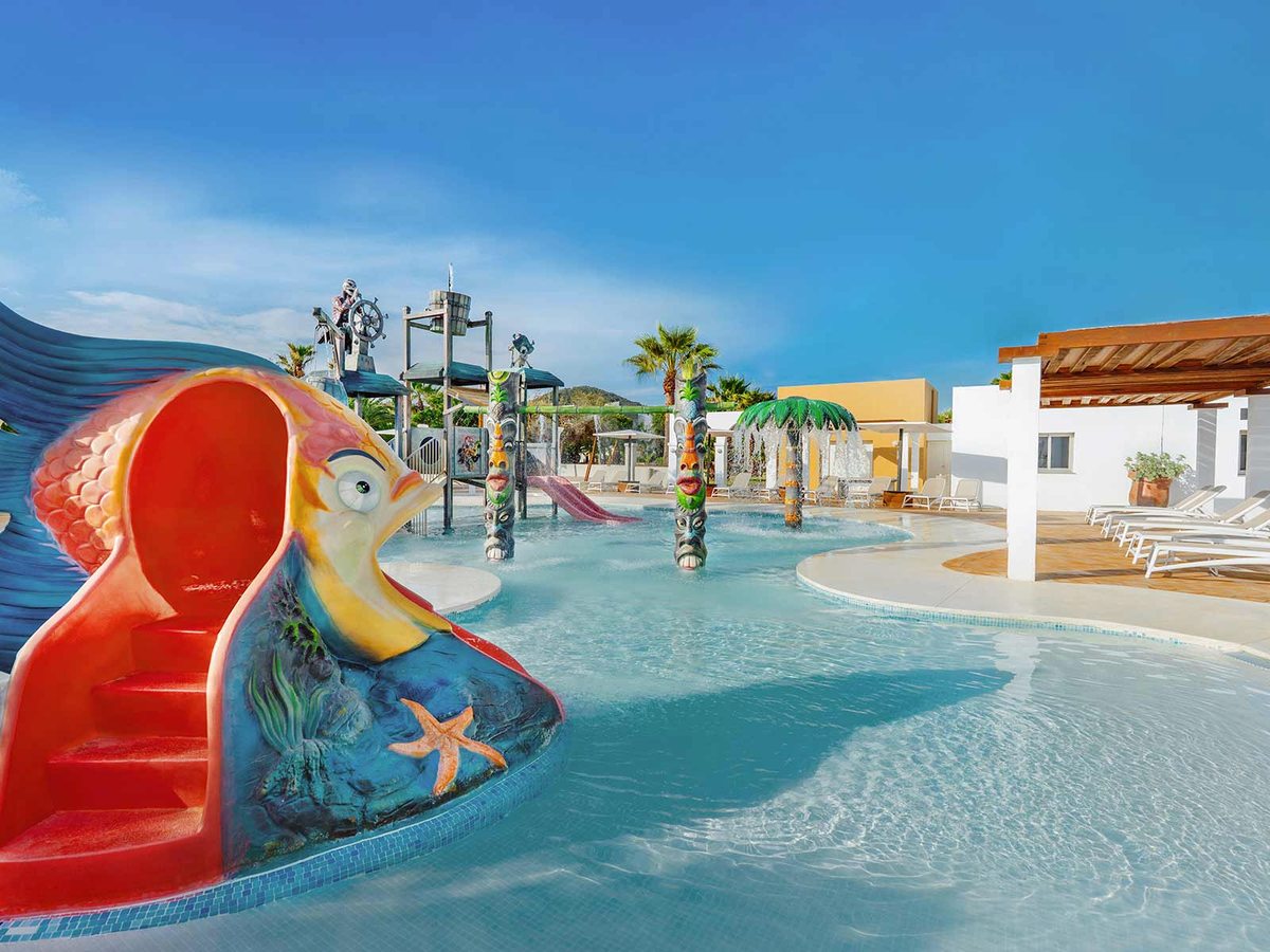 Children's water park with a colourful fish-shaped slide, a tipping-bucket play tower and a shallow pool under a blue sky.