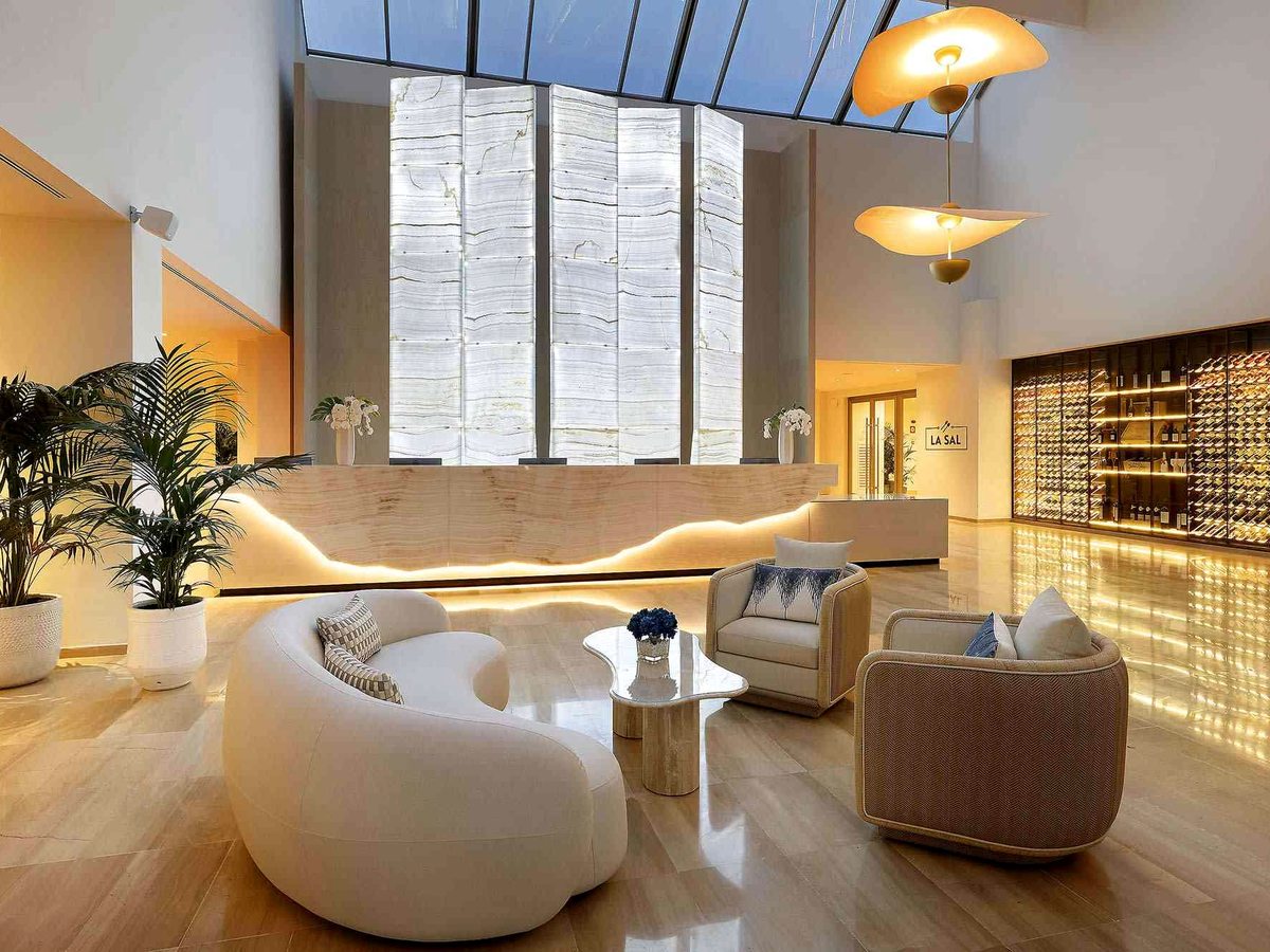 Open lobby lounge with a curved cream sofa, low armchairs and a backlit stone feature wall under a high glazed roof.