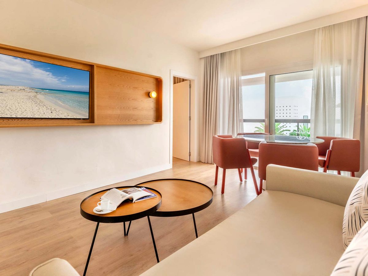 Suite living room with a sofa bed, coffee tables and a dining table with red chairs, a wall-mounted TV and balcony doors.