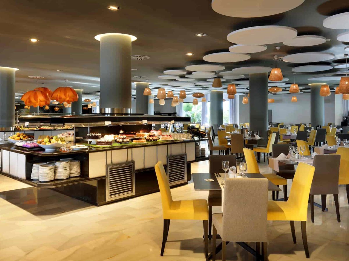 Large buffet restaurant with self-service food islands under round ceiling lights and rows of tables with yellow chairs.