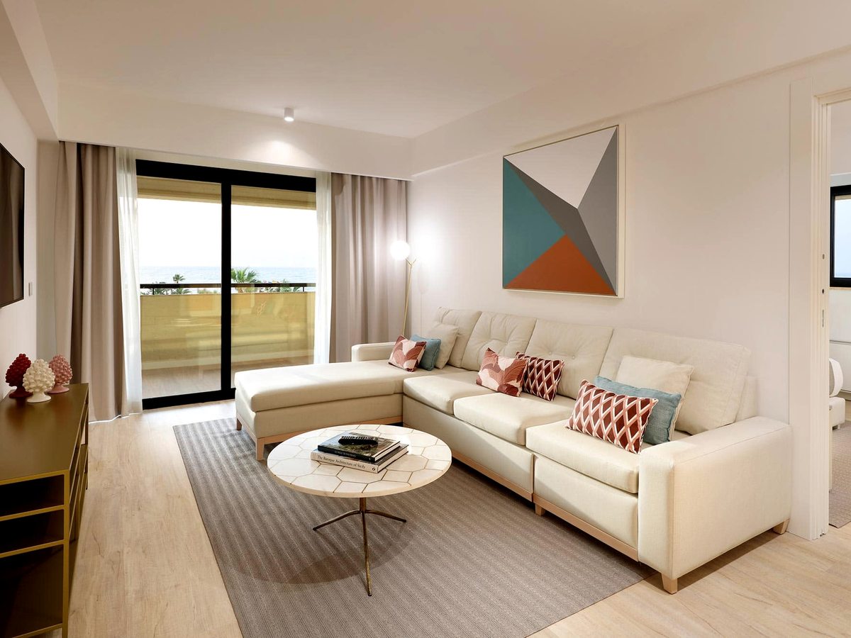 A suite sitting room with an L-shaped cream sofa, a round coffee table, a wall-mounted TV and a geometric artwork above.