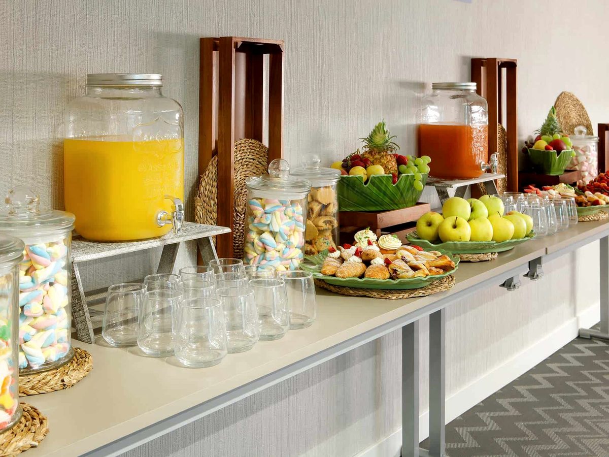 A buffet counter set with tall glass dispensers of juice, stacked tumblers, jars of sweets and platters of pastries and fruit under warm lighting.