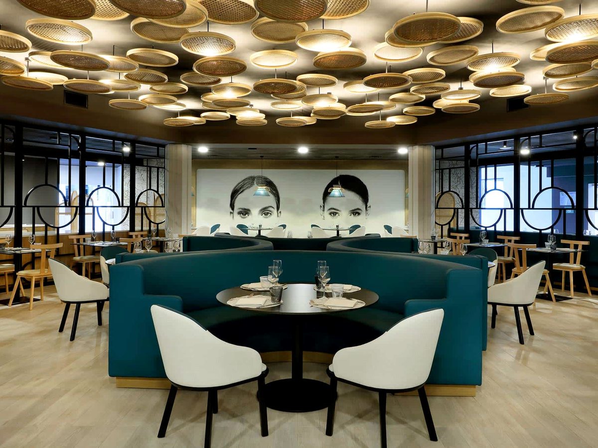 A restaurant dining room with a curved teal banquette booth, round tables laid with glassware, patterned chairs and a coffered wooden ceiling.