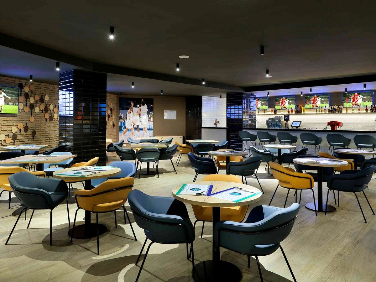 A sports bar with wall-mounted screens, round tables and blue and mustard tub chairs on a patterned floor, with a bar counter along one side.