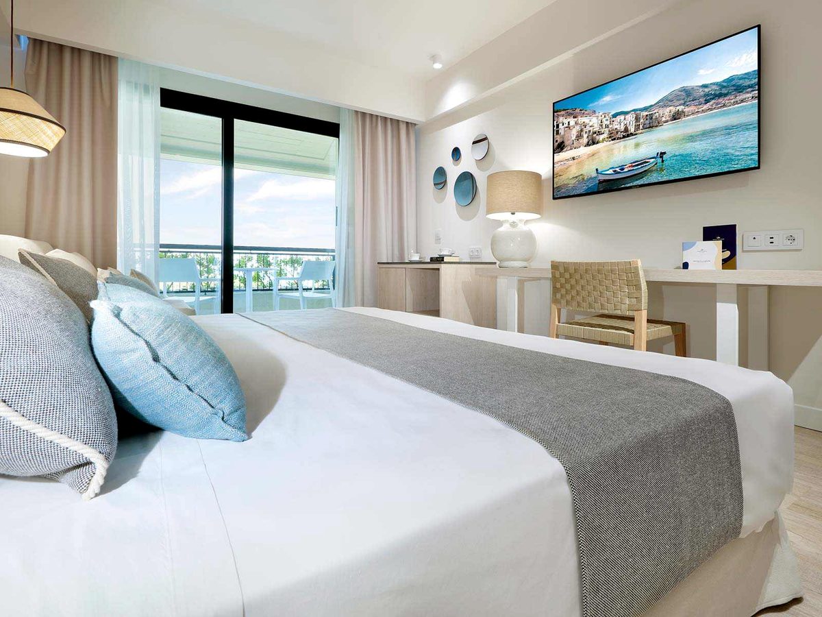 A double room with a large bed and blue scatter cushions, a desk and chair, a wall-mounted TV and sliding glass doors to a balcony.