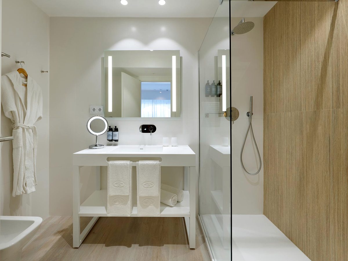 A modern bathroom with a white double vanity and illuminated mirrors, folded towels and bathrobes, and a walk-in rainfall shower behind glass.