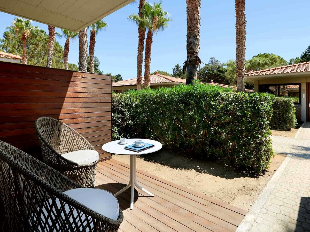 A single-storey bungalow with a private wooden-deck terrace, cane armchairs and a low table, set behind a clipped hedge with palms and terracotta-roofed bungalows beyond.