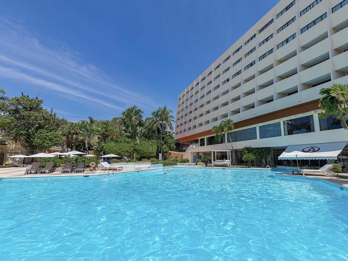 A large white multi-storey hotel building behind a big curving blue swimming pool ringed by sun loungers, parasols and palms under a clear blue sky.