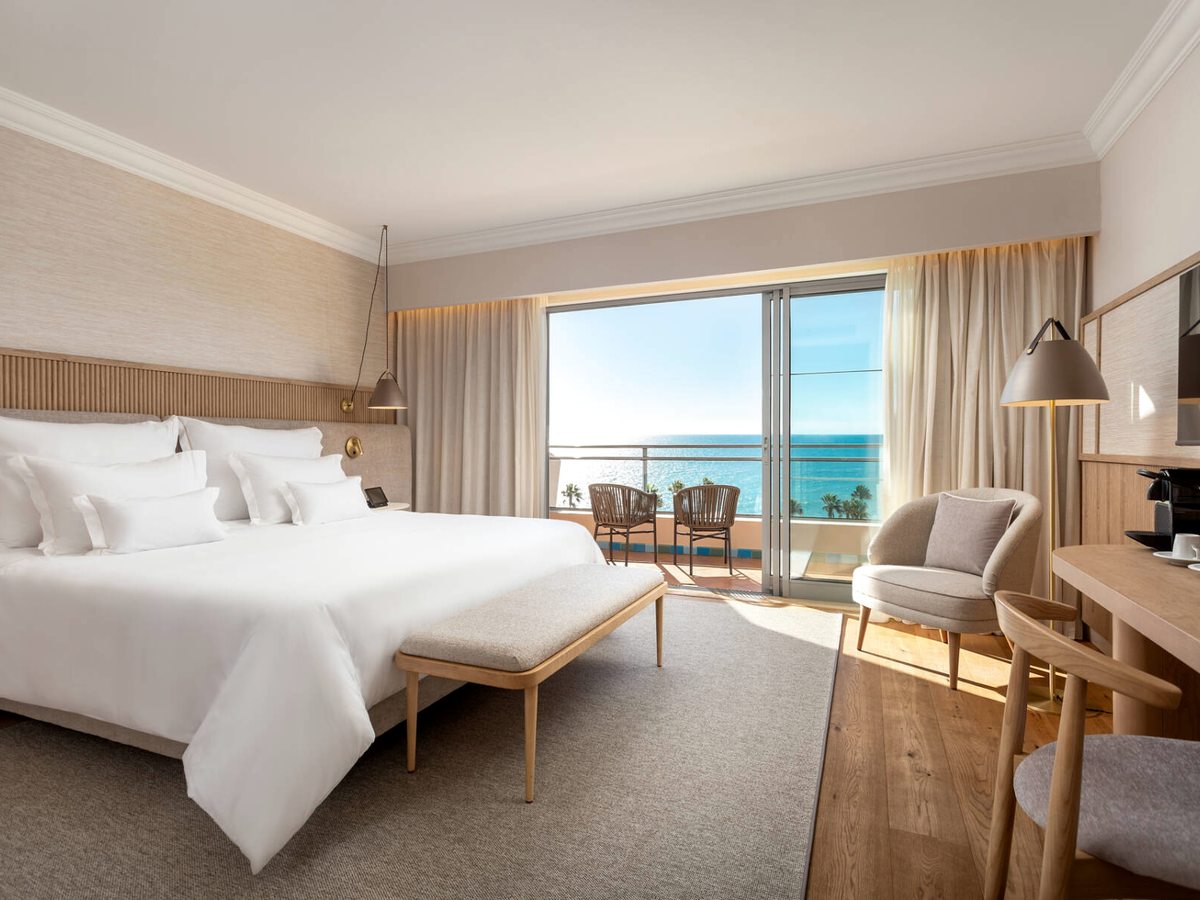 A contemporary room with a king bed in white linen, a padded bench, and a balcony with two chairs facing the sea.