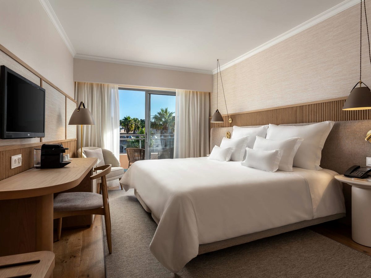 A renovated room with a double bed, a writing desk with a coffee machine and a balcony framing palms and gardens.