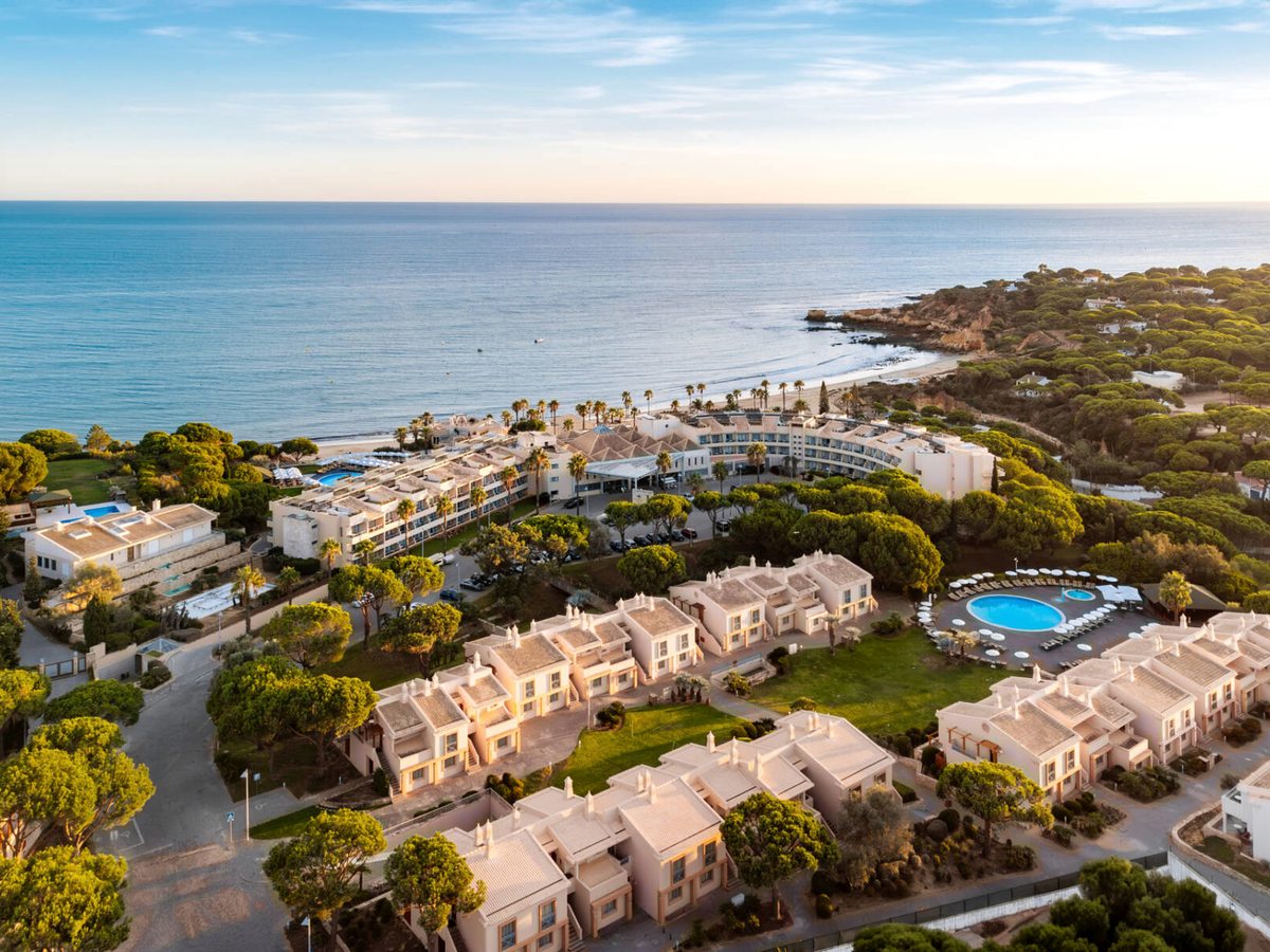An aerial view of white resort buildings and gardens along a green, cliff-backed coastline.