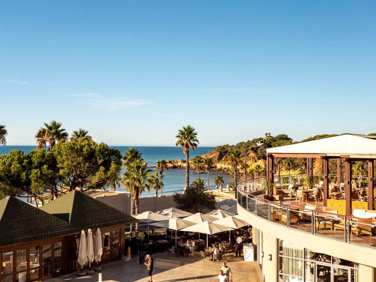 Outdoor dining terraces with cream parasols and palm trees on the clifftop above the sea.