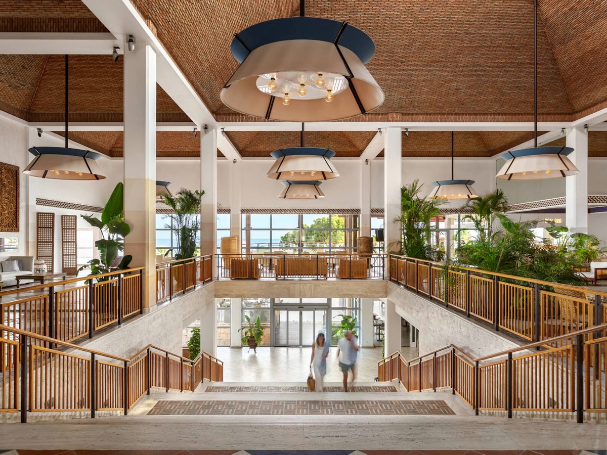 A bright multi-level atrium with wooden staircases and railings, tall plants and a woven timber ceiling.