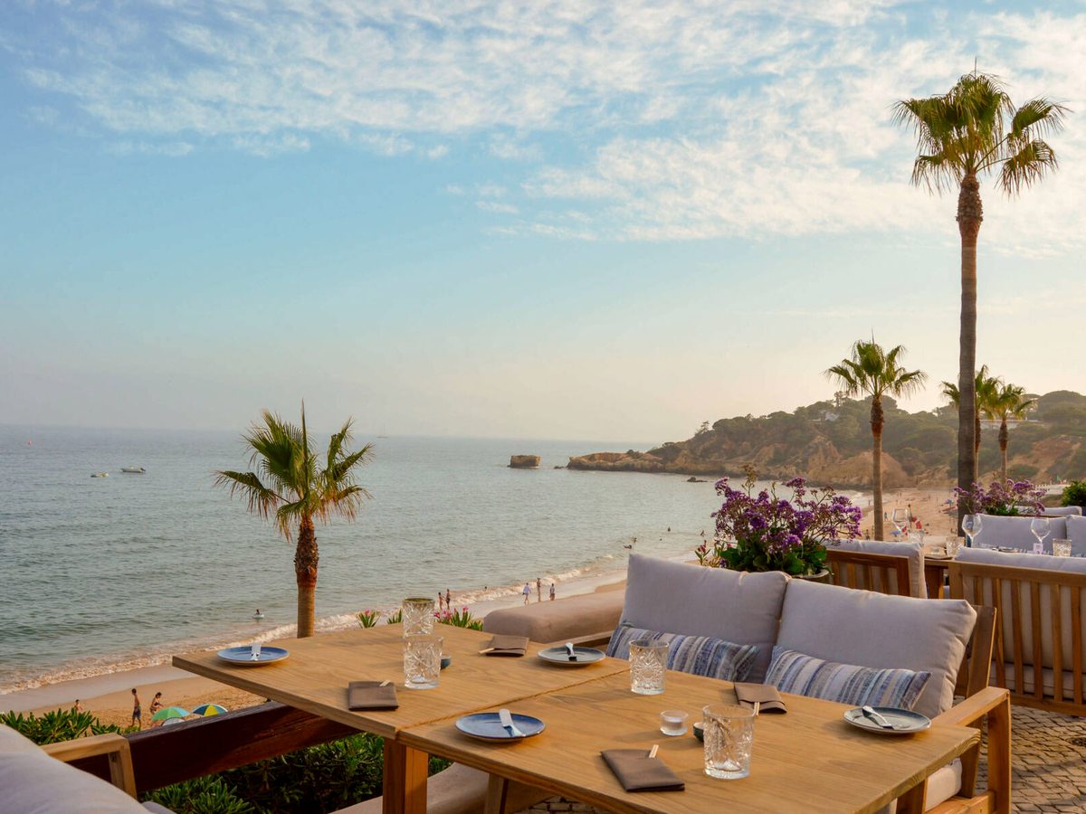 A wooden dining table laid with glasses and napkins on a terrace, palms and the sandy cove of Santa Eulália beach beyond.