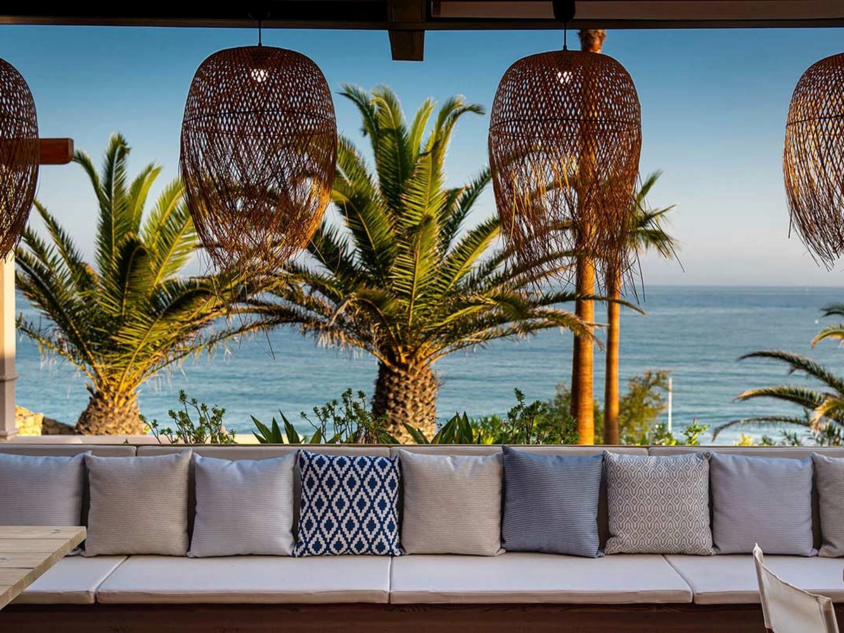 A long cushioned banquette under woven pendant lamps, with palms and the sea framed beyond.