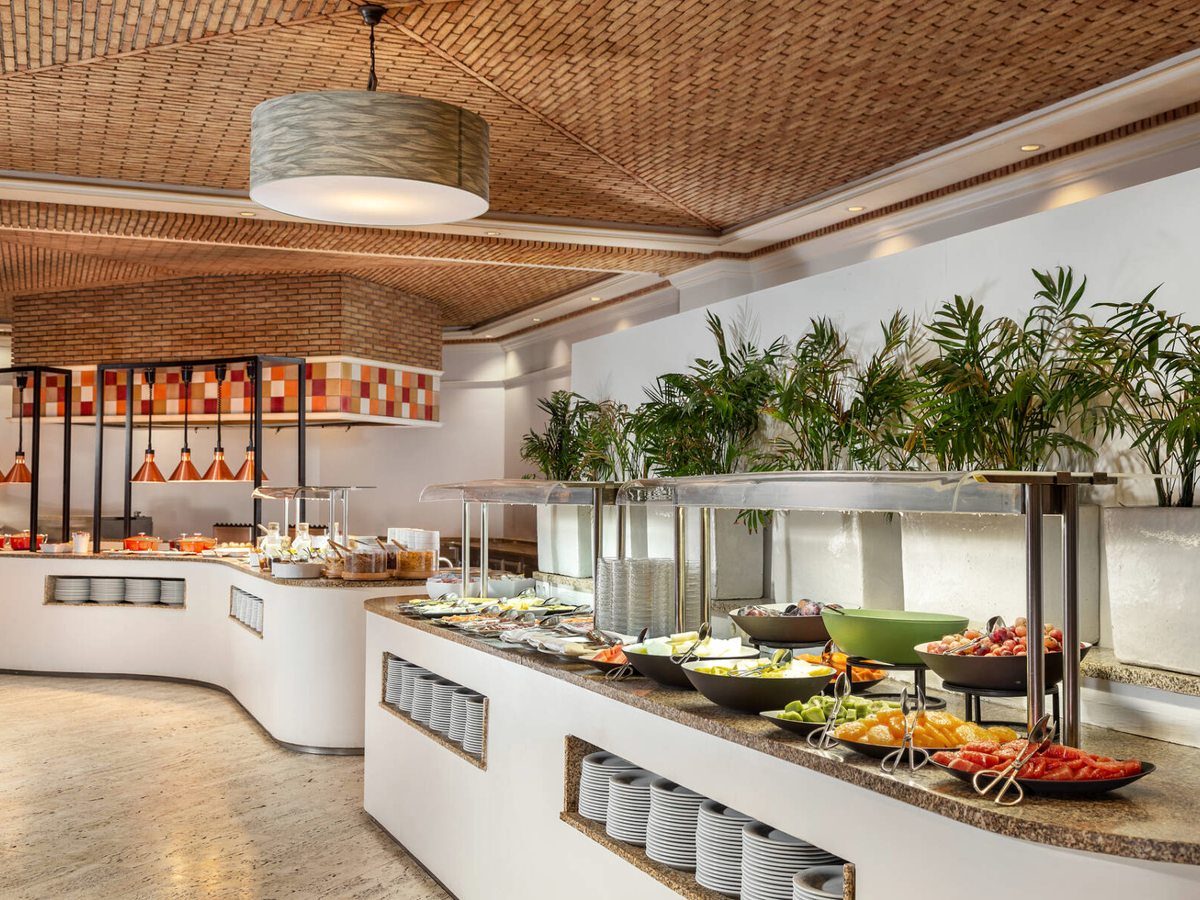 A curved buffet counter with bowls of fruit and salad under glass guards, beneath a woven ceiling and copper heat lamps.