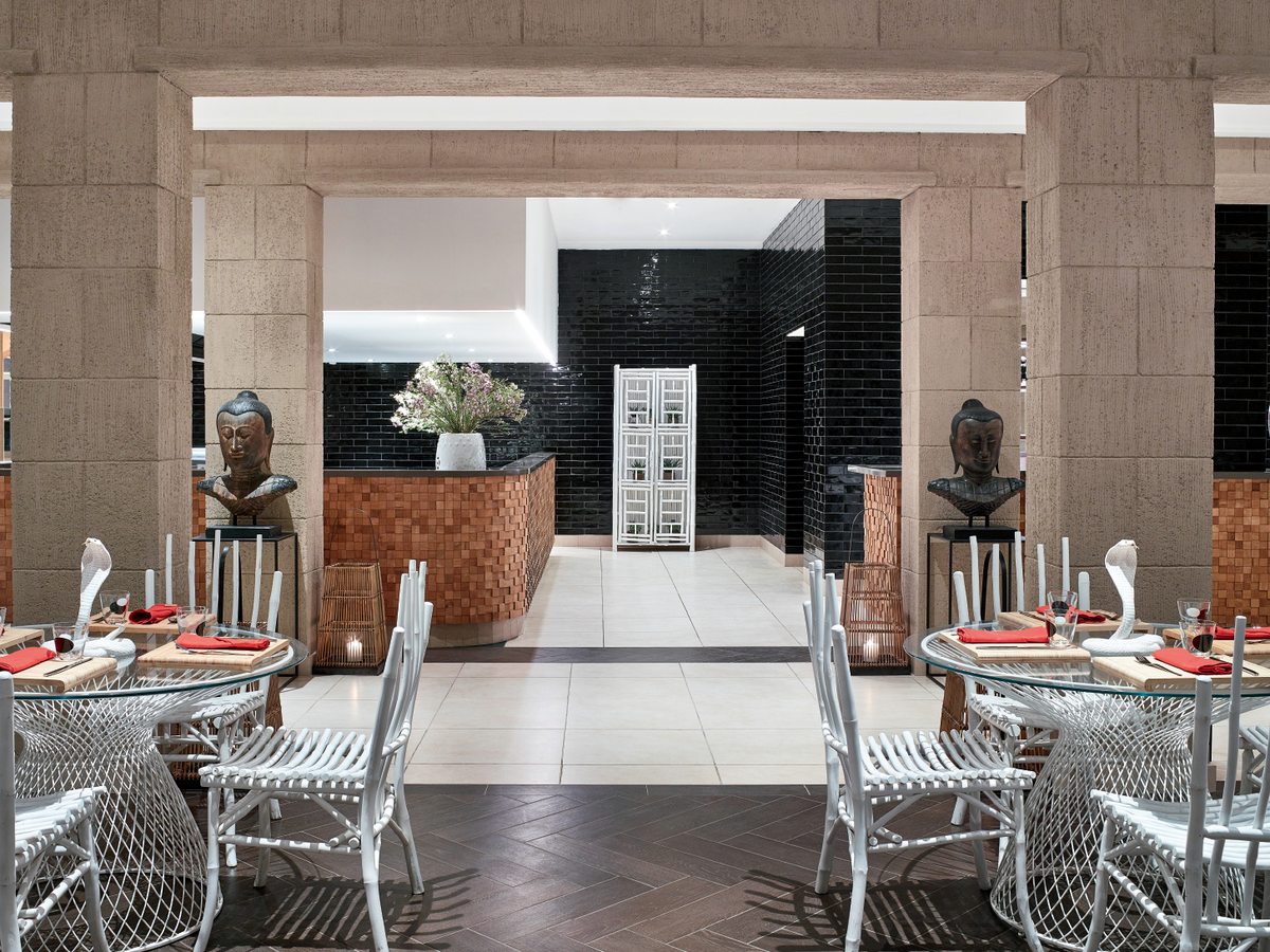 The indoor dining room at Asiana, with white rattan chairs, glass-topped tables set with red napkins and carved Buddha busts flanking the entrance.